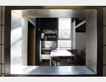 Blick durch eine Edelstahl-Durchreiche in eine industriell eingerichtete Küche mit Fliesenwänden, Edelstahl-Arbeitsflächen, Spüle und Regalen mit Reinigungsmitteln. Im Hintergrund ist ein Fenster mit Gitterstäben sichtbar.