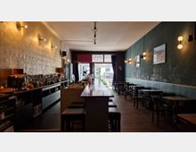 Langer, schmaler Innenraum einer Bar mit dunkler Holzeinrichtung. Links steht eine Theke mit Flaschenauslage, rechts befinden sich Tische und Stühle an einer dunkelgrünen Wand mit dekorativen Lampen und Bildern; im Hintergrund ist ein Fenster mit Blick nach draußen erkennbar.