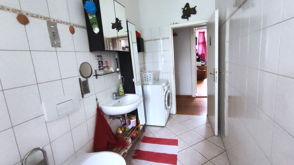 Narrow bathroom with white tiled walls and a diagonal-tile floor, featuring a sink with mirror and shelves, wall cabinets, and a toilet in the foreground. A washing machine sits near an open door leading to a hallway, with a red-and-white striped rug on the floor.