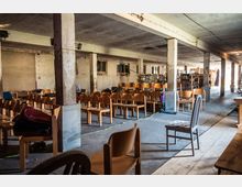 Ein großer, schlichter Raum mit rohem Betonboden und sichtbaren Säulen; im Vordergrund stehen zahlreiche hölzerne Stühle in Reihen, während im Hintergrund weitere Möbel gegen die Wände gelagert sind. Der Raum wirkt unrenoviert mit unverputzten Wänden und spärlichem Licht, das durch kleine Fenster einfällt.