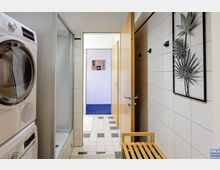 Waschraum mit weiß gefliesten Wänden, einer Waschmaschine und einem Trockner auf der linken Seite. Rechts sind Wandhaken und eine dekorative Metall-Wandverzierung mit Blattmotiven zu sehen, daneben steht eine Holzbank. Eine offene Tür führt in einen angrenzenden Raum mit Teppichboden.