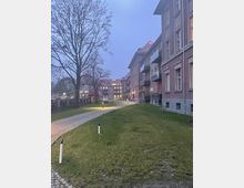 Außenbereich eines Wohnkomplexes bei Dämmerung, mit gepflastertem Gehweg, Rasenflächen und beleuchteten Wegmarkierungen. Mehrstöckige Gebäude aus Backstein mit Balkons auf der rechten Seite und einem großen, kahlen Baum links im Bild.