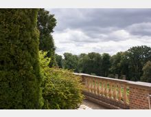 Eine halbhohe Steinbalustrade mit Ziersäulen grenzt an eine Terrasse, die von üppig grünem Buschwerk und hohen Bäumen umgeben ist. Der Himmel ist bewölkt, und im Hintergrund ist eine bewaldete Landschaft zu sehen.