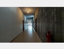 Ein langer, schmaler Flur in einem modernen Gebäude mit glatten Wänden, die links weiß und rechts aus Sichtbeton sind. Der Boden ist beige, und am Ende des Flurs befindet sich eine verglaste Tür mit Tageslichteinfall; rechts im Vordergrund steht ein roter Feuerlöscher.