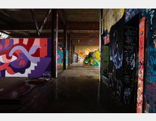 Innenraum einer verlassenen Industriehalle mit Betonboden, Stahlträgern und niedriger Decke, durchzogen von einem Gang. Wände und Pfeiler sind dicht mit großformatigen Graffiti und Street-Art-Motiven in kräftigen Farben bedeckt.