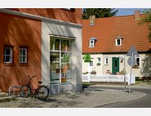 Straßenszene in einer Kleinstadt mit rot-orange verputztem Haus links und einem kleinen, hell gestrichenen Haus mit roten Ziegeln und weißem Zaun rechts. Ein Fahrrad steht vor dem Schaufenster eines Geschäfts, und Straßenschilder sind im Vordergrund zu sehen.