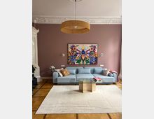 Elegantes Wohnzimmer mit hoher Stuckdecke, mauvefarbener Wand und großer runder Hängelampe aus Rattan. Vor der Wand steht ein hellblaues Sofa unter einem farbenfrohen Gemälde, davor ein gläserner Couchtisch auf einem hellen Teppich über Parkettboden.