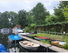 Kleiner Bootsanleger an einem ruhigen Fluss mit mehreren kleinen Booten, darunter Tretboote und Ruderboote, sowie einigen größeren Motorbooten. Im Hintergrund sind ein rotes Haus mit Schrägdach, grüne Bäume und eine dichte Hecke zu sehen.