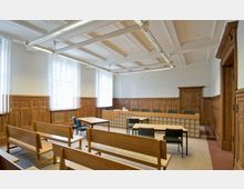 Ein heller Gerichtssaal mit Holztäfelung an den Wänden, einem verzierten Kassettendeckensystem und großen Fenstern mit vertikalen Lamellen. Im Raum stehen Holzbänke, Tische und Stühle sowie ein erhöhtes Pultbereich für Richter oder Redner.