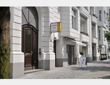 Fassade eines mehrstöckigen Altbaus mit hell gestrichener Putzfassade, großen Fenstern und Balkonen. Links im Vordergrund befindet sich ein großes, dunkles Holztor, darüber ein Schild mit der Aufschrift „Pension Funk“. Rechts davon ist ein Geschäftseingang mit Schaufenstern und einem aufgestellten Aufsteller zu sehen.