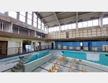 Ehemaliges Hallenbad mit leerem, verfallenem Schwimmbecken. Die Wände sind teils mit blauen und türkisfarbenen Fliesen verkleidet, umgeben von Umkleidekabinen und einer Galerie mit Geländer; im Becken stehen Stühle.