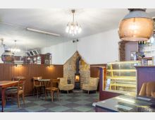 Gemütliches Café mit Holzwänden und einer Wand aus Sichtstein, in der ein kleiner Kamin eingebaut ist. Im Raum stehen verschiedene Holztische und Stühle, eine Glasvitrine sowie eine Bar mit Getränken in Regalen.