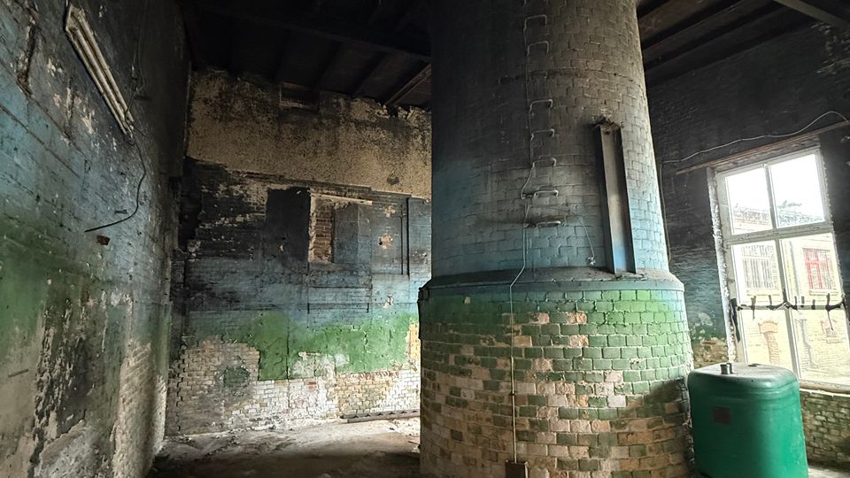 Innenraum eines alten Industriegebäudes mit rußgeschwärzten Ziegelwänden und freiliegenden Deckenbalken. In der Mitte steht ein großer zylindrischer Schornstein- oder Ofenschacht aus Ziegeln, daneben ein grüner Kunststoffbehälter am Fenster.