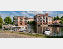 Ziegelgebäude mit modernen Balkonen, die an einem Flussufer stehen und durch einen bogenförmigen Übergang verbunden sind, mit einem kleinen Bootsanleger im Vordergrund und Booten auf dem Wasser. Im Hintergrund sind Bäume und ein weiteres Gebäude zu sehen, unter einem Himmel mit leichten Wolken.