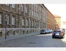 Gepflasterte Straße mit parkenden Autos entlang zweier mehrstöckiger Gebäude: links ein graues Gebäude mit klassischen Fensterrahmen und rechts ein roter Ziegelbau. Laternen und Gehwege verlaufen beidseitig entlang der Gebäude.