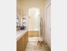 Modernes Badezimmer mit hellen Fliesen, einem Waschtisch aus Holz mit einer eingelassenen weißen Waschschüssel und einem Spiegel mit Leuchtrahmen. Im Hintergrund eine freistehende, beigefarbene Badewanne vor einer halbhohen Wand und einer Duschnische mit Glaswand, daneben ein Handtuchhaken mit aufgehängtem Bademantel.