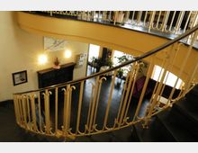 Innenraum mit einer geschwungenen Wendeltreppe und einem dekorativen Geländer, das in ein offenes Stockwerk mit schwarzen Fliesen führt. Der Bereich enthält Pflanzen, gepolsterte Sitzmöbel und Wandleuchten, die warmes Licht ausstrahlen.