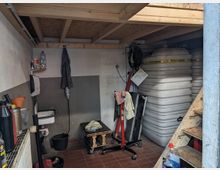 Ein kleiner, funktionaler Lager- oder Werkstattraum mit Holzbalkendecke, Wassertank, Waschbecken, Werkzeugen und Regalen. Der Boden besteht aus roten Fliesen, und auf der rechten Seite führt eine Holztreppe nach oben.