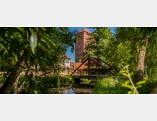 Blick durch dichte Uferbepflanzung auf einen kleinen Bach mit einer roten Stahl-Fußgängerbrücke. Im Hintergrund steht ein hoher Backstein-Burgturm mit angrenzenden Gebäuden, umgeben von Bäumen unter blauem Himmel.