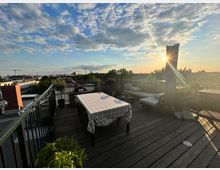 Dachterrasse mit Holzboden, auf der ein Tisch mit gestreifter Tischdecke und Stühlen sowie eine Sitzecke mit Polstermöbeln steht. Im Hintergrund ist eine Stadtlandschaft mit grünem Baumkronenbereich und Sonnenuntergang zu sehen, der Himmel ist teils bewölkt.