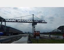 Industriehafen mit einem großen, stählernen Verladekran, der sich über eine gepflasterte Straße erstreckt. Im Hintergrund sind Lagerhallen, Schienen und ein schmaler Wasserkanal zu sehen, eingerahmt von einer bewölkten Himmelsszenerie.