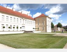 Weiße barocke Schlossfassade mit rotem Ziegeldach, großen rechteckigen Fenstern und dekorativem Eingangsbereich, umgeben von gepflegtem Rasen und einzelnen Topfbäumen. Im Hintergrund sind weitere historische Gebäude, Bäume und ein teils bewölkter Himmel zu sehen.