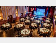 Festlich dekorierter Veranstaltungssaal mit runden Tischen, die mit weißen Tischdecken, grünen Servietten und Kerzenleuchtern geschmückt sind. Im Hintergrund eine Bühne mit rotem Vorhang, bunter Beleuchtung und dekorativen Elementen.