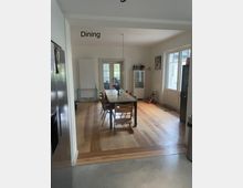 Essbereich in einem modernen Innenraum mit Holzboden, einem rechteckigen Esstisch mit Holzstühlen und einer Pendelleuchte über dem Tisch. Im Hintergrund stehen eine weiße Glasvitrine, Doppeltüren mit Glaseinsatz und ein großes Fenster, das Tageslicht hereinlässt.