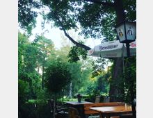 Ein Biergarten im Freien, umgeben von hohen Bäumen und dichter Vegetation. Holztische und Bänke stehen unter Sonnenschirmen mit der Aufschrift „Benediktiner Weißbier“, neben einer Laterne.