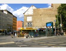 Straßenszene am Rosenthaler Platz in Berlin mit U-Bahn-Eingang, einer Bäckerei mit gelbem Vordach, graffitiverzierter Backsteinwand und umliegenden mehrstöckigen Gebäuden. Im Vordergrund sind Gehwege, Schienen, Fahrräder und Passanten zu sehen.