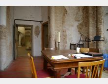 Ein Raum mit unverputzten Backsteinwänden und sichtbaren Abnutzungsspuren, eingerichtet mit einem Holztisch, mehreren Stühlen und Kerzenständern; im Hintergrund führt eine Tür in einen Bereich mit einer Wendeltreppe und weiteren historischen Elementen.