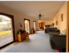 Langer Wohnraum mit großen, bogenförmigen Fenstern, die auf eine Terrasse mit gepflastertem Boden und Rasen führen. Der Raum enthält eine Sitzecke mit grauen Sofas, ein hölzernes Sideboard, Regale und eine Bar mit Stühlen im Hintergrund.