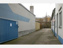 Schmaler gepflasterter Weg zwischen Gebäuden mit schlichten, hellen Fassaden; links befindet sich ein blaues Metalltor und rechts ein Hinweisschild "Schritt fahren". Im Hintergrund sind Bäume und ein größeres Industrie- oder Bürogebäude zu sehen.