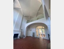 Heller Innenraum mit hoher, gewölbter Decke, roten Fliesenböden und großen Türbögen. Links ein Kamin und rechts eine Glastür, die zu einem sonnigen Raum mit großen Fenstern führt.