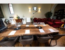 Ein stilvoll eingerichteter Raum mit einem großen Holztisch in der Mitte, der von gepolsterten Stühlen umgeben ist. Im Hintergrund befinden sich ein rotes Sofa, dekorative Elemente wie Kerzenhalter, Pflanzen und eine Bar, die eine gemütliche Atmosphäre schaffen.