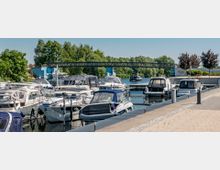 Jachthafen mit mehreren festgemachten Motorbooten entlang eines Stegs, umgeben von Bäumen und Grünflächen. Im Hintergrund führt eine schmale Fußgängerbrücke über das Wasser.