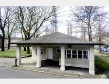 Kleines einstöckiges Gebäude mit Putzfassade und Walmdach, das von Säulen gestützt wird. Das Gebäude steht in einer parkähnlichen Umgebung mit kahlen Bäumen und Rasenflächen, während im Hintergrund eine Straße und geparkte Autos zu sehen sind.