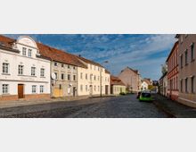 Straßenszene in einer Kleinstadt mit gepflasterter Fahrbahn, gesäumt von zweigeschossigen Wohnhäusern mit verputzten Fassaden und roten Ziegeldächern. Im Vordergrund stehen geparkte Autos, und der Himmel ist leicht bewölkt.