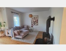 Helles, modern eingerichtetes Wohnzimmer mit L-förmigem Stoffsofa auf hellem Teppich, kleinem Beistelltisch und großem Blumenbild an der Wand. Links sorgt ein Fenster mit weißen Vorhängen für Tageslicht, rechts steht ein schwarzer Kaminofen mit Ofenrohr auf Holzboden.