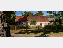 Einfache ländliche Hausfassade mit einem roten Ziegeldach, umgeben von Bäumen und einem Metallzaun im Vordergrund. Der Garten ist teilweise gepflegt, mit einem einzelnen kleinen Baum in der Mitte.