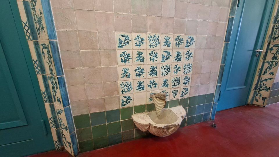 Innenraum mit gefliester Wand in Beige und Grün, in der Mitte ein kleines Wandbecken aus Stein unter einem Feld blau-weiß gemusterter Kacheln. Links und rechts rahmen türkisfarbene Türen und dekorative Kachelstreifen den Bereich, der Boden ist rot.