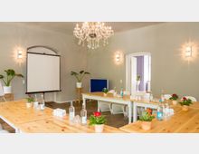 Heller Seminarraum mit Holztischen, darauf Wasserflaschen, Gläser und Topfpflanzen. Im Hintergrund befinden sich ein Kronleuchter, eine Leinwand, Zimmerpflanzen und eine offene Tür, die in einen weiteren Raum führt.