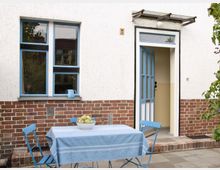 Außenansicht eines Hauses mit weißer Fassade und rotem Klinkersockel. Im Vordergrund steht ein blauer Klapptisch mit Tischdecke und einer Schale mit Obst, daneben zwei passende blaue Stühle; rechts befindet sich eine blaue Eingangstür, links ein Fenster mit hellblauem Rahmen.