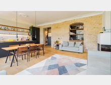 Wohnzimmer mit offener Küche in einem modernen, gemütlich eingerichteten Apartment. Sichtbare Backsteinwand, Holzboden, Esstisch mit Stühlen, graues Sofa, Regale und eine Küchenzeile mit schwarzen Elementen dominieren den Raum; im Vordergrund ein geometrisch gemusterter Teppich.