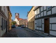 Pflastersteinstraße in einer Altstadt mit Fachwerkhäusern und gelb gestrichenen Gebäuden; im Hintergrund ein Kirchturm aus rotem Backstein mit weißem Dachelement. Autos parken entlang der Straße, und Laternen säumen die Gasse.