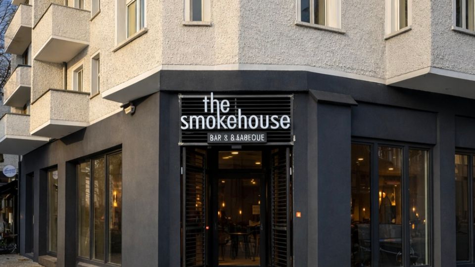 Ecke eines mehrstöckigen Wohn- und Geschäftshauses mit heller Putzfassade und Balkonen; im dunklen Erdgeschoss befindet sich ein Restaurant mit großen Schaufenstern und einem Eingang unter dem Schild „the smokehouse“. Vor dem Gebäude liegt ein breiter Gehweg mit Pflaster- und Steinplattenbelag.