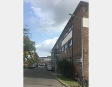 Mehrstöckiges Backsteingebäude mit modernen Fenstern und einer außenliegenden Metalltreppe, das sich an einer asphaltierten Straße mit parkenden Autos befindet. Im Hintergrund sind weitere Gebäude im ähnlichen Stil zu sehen, teils umgeben von Bäumen unter leicht bewölktem Himmel.