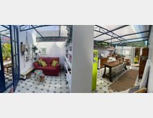 Ein Wohnzimmer und eine Küche in einem lichtdurchfluteten Raum mit verglastem Dach. Das Wohnzimmer enthält ein rotes Sofa, einen kleinen Tisch und Hängepflanzen, während die Küche mit einer langen Holztischgruppe, einer grünen Retro-Kühlschranktür und gemusterten Fliesen dekoriert ist.