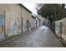 Eine gepflasterte Gasse mit alten, von Graffiti bedeckten Hausfassaden auf der linken Seite und einer Natursteinmauer auf der rechten Seite. Kahle Bäume und eine Straßenlaterne säumen den Weg, während im Hintergrund Personen zu Fuß zu sehen sind.