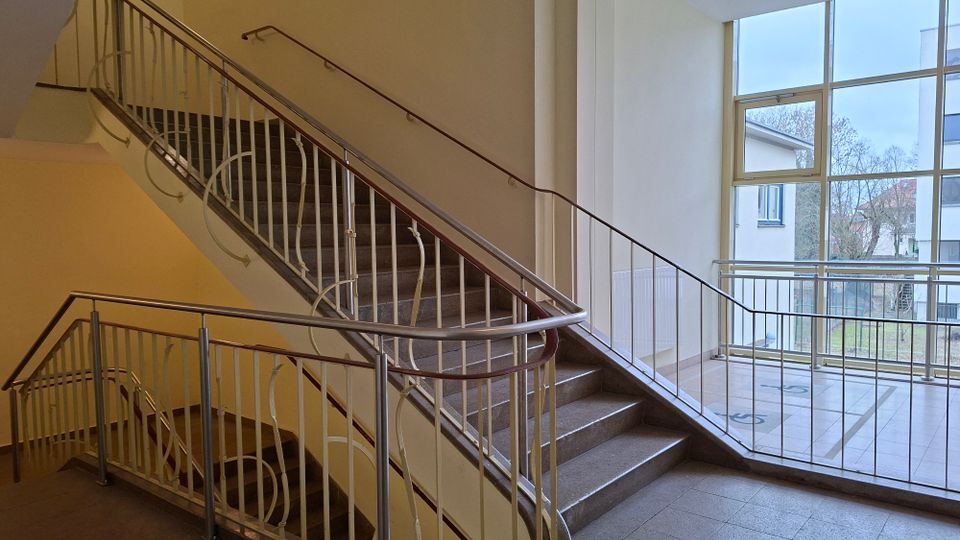Innenansicht eines Treppenhauses mit zweiläufiger Treppe, grauen Stein- bzw. Betontreppen und weißen Metallgeländern mit Handlauf. Rechts sorgt ein großes, mehrteiliges Fenster für Tageslicht und bietet Blick auf umliegende Gebäude und Bäume.